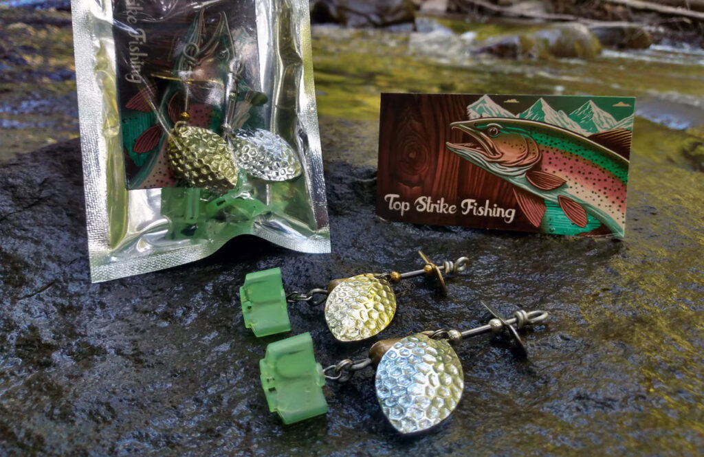 The best trout fishing spinners on a rock with product packaging, business cards, and hook caps. Creek water in the background.