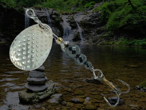 An inline spinner made for trout with silver and black components. Waterfall, creek and greenery in the background.