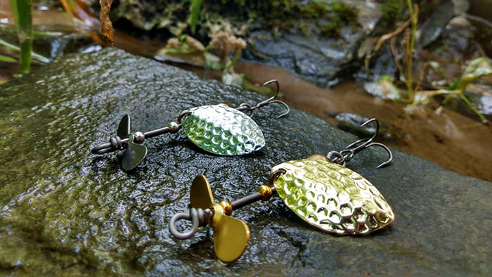 Two propeller fishing lures on a large stone that is half wet, with vegetation in the background.