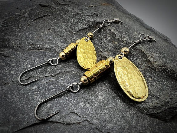 A single-hook trout spinner with a hammered blade, polished gold components. Photographed on a dark rock.