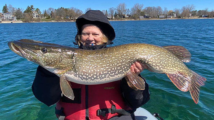 Barb Labignan of Canadian Sportsfishing holds a trophy pike. Beautiful lake scene in the background.