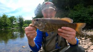 Angler holds smallmouth bass with wooded creek in the background.