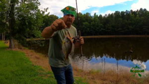 Justin Peden of Spirit of the Outdoors holds a bluegill by the lip with a farm pond and forest in the background.