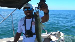 Anthony of KeepFishing on YouTube holds a small walleye up to the camera with fish lip grips. Distant lake view in the background.