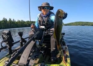 OldLadyAngler of YouTube holds a largemouth bass up to the camera by its lip. Maine mountain pond in the background.