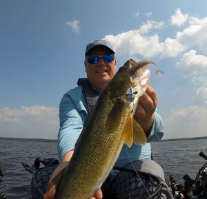 FisherMN of YouTube holds a walleye with two hands while sitting on a kayak in a lake. Top Strike Fishing spinner in the walleye's mouth.