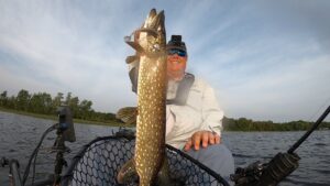 FisherMN of YouTube holds a northern pike vertically by its rear gill plate while sitting in a kayak on a lake. Top Strike Fishing spinner hangs from the pike's mouth.