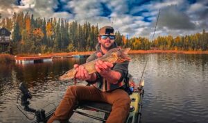 Chris Lindner (907shots) holds a pike while sitting in a kayak on the water. Autumn colors in the background.